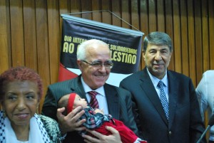 Emir Mourad, ladeado por Leci Brandão e Ibrahim Alzeben, segura a filha recém-nascida de uma refugiada palestina / Foto: Hasan Rabee